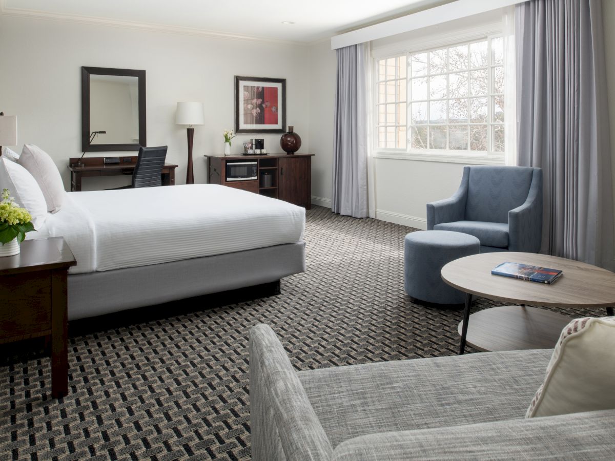 A modern hotel room features a bed, a desk with a mirror, an armchair with a table, and a sofa, all in a light, relaxing decor.
