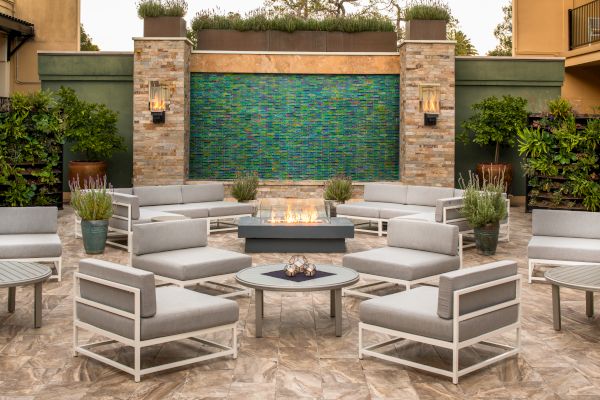 An outdoor seating area with modern lounge chairs, a central fire pit, a water feature on a tiled wall, and surrounding plants.