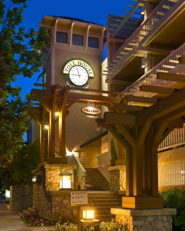 The image shows a building with illuminated wooden beams, stone accents, and a clock tower at the front, under a deep blue evening sky.